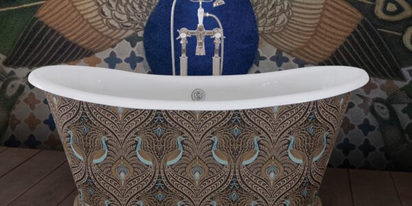 The image showcases a luxurious freestanding bathtub with a bold maximalist design, set against a similarly ornate background. The exterior of the tub is adorned with a peacock feather pattern, rendered in a palette of rich browns, golds, and teals. This intricate pattern gives the tub an opulent and bespoke appearance. The bathtub fixtures are traditional in style with a chrome finish, adding a touch of timeless elegance to the modern expression of maximalism. Behind the bathtub, a peacock-themed wall mural complements the tub's design, featuring an expanded tail in a display of vibrant green, blue, and gold hues, set against a deep blue background. The floor is a wooden texture, providing a warm, natural contrast to the cool tones of the tub and wall. The overall effect is one of lavishness and artistic flair, making a striking statement in this maximalist bathroom setting.