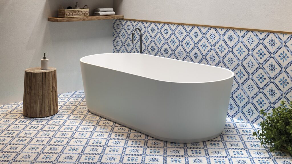 The image shows a modern bathroom setting with a focal point on a standalone bathtub. The bathtub is white with a smooth, curved design, likely made of acrylic or porcelain, and it's positioned in the centre of the room. Behind the tub, there is a section of the wall decorated with blue and white patterned tiles that add a traditional and somewhat Mediterranean touch to the space. The floor is adorned with matching tiles, creating a cohesive and vibrant aesthetic. To the left of the tub, there's a wooden stool that holds a dispenser bottle, possibly soap or lotion. Above the bathtub, a wooden shelf stretches across the wall, holding neatly stacked towels and some bathroom essentials in a wicker basket. On the right side, there is a green plant that introduces a natural element and a pop of colour to the scene. The overall look of the bathroom is clean, serene, and stylish, with a blend of modern fixtures and classic decorative tiles.