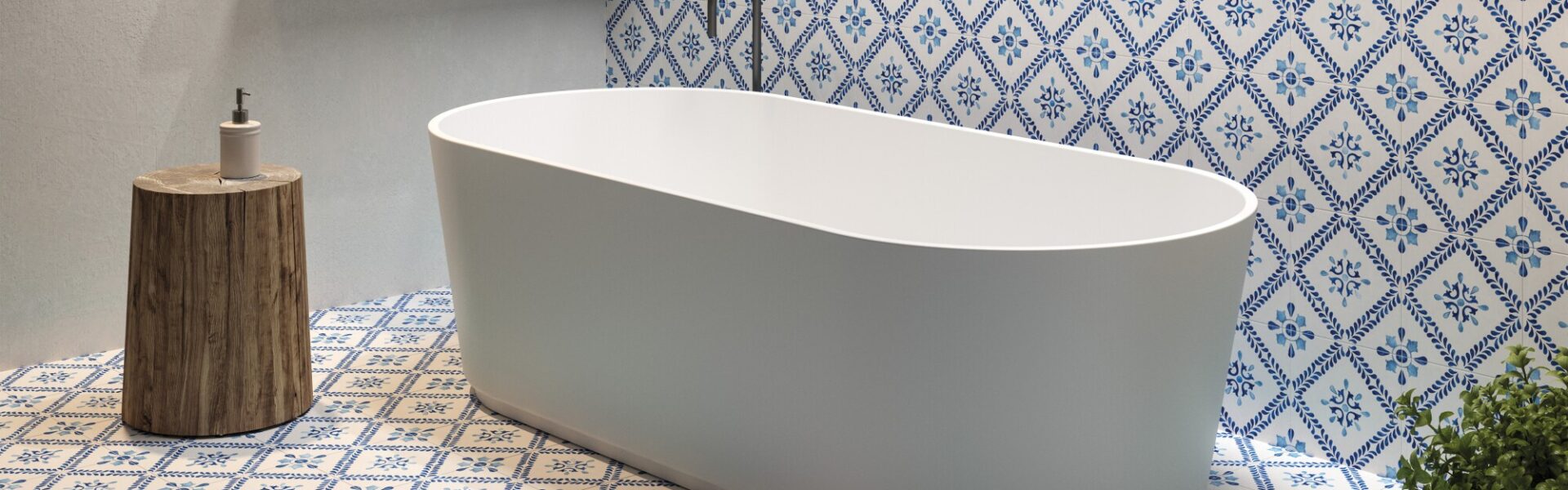 The image shows a modern bathroom setting with a focal point on a standalone bathtub. The bathtub is white with a smooth, curved design, likely made of acrylic or porcelain, and it's positioned in the centre of the room. Behind the tub, there is a section of the wall decorated with blue and white patterned tiles that add a traditional and somewhat Mediterranean touch to the space. The floor is adorned with matching tiles, creating a cohesive and vibrant aesthetic. To the left of the tub, there's a wooden stool that holds a dispenser bottle, possibly soap or lotion. Above the bathtub, a wooden shelf stretches across the wall, holding neatly stacked towels and some bathroom essentials in a wicker basket. On the right side, there is a green plant that introduces a natural element and a pop of colour to the scene. The overall look of the bathroom is clean, serene, and stylish, with a blend of modern fixtures and classic decorative tiles.