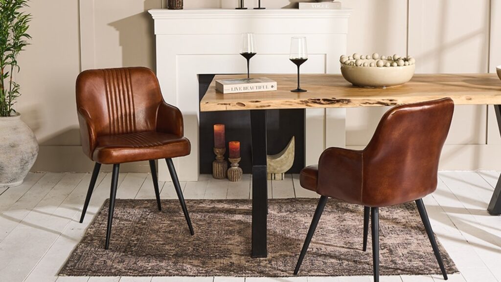 A contemporary dining room featuring a rustic wooden table with live edges, flanked by two brown leather chairs with black metal legs. The table is set against a muted beige wall with a white fireplace mantel. On the table, there's a decorative bowl filled with spheres, a book titled 'Live Beautiful', and two black-stemmed wine glasses. The scene is completed by a textured dark rug underneath and a large potted plant to the left, adding a touch of greenery to the warm-toned space.