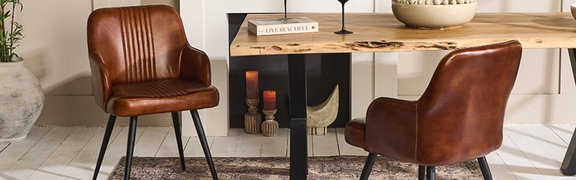 A contemporary dining room featuring a rustic wooden table with live edges, flanked by two brown leather chairs with black metal legs. The table is set against a muted beige wall with a white fireplace mantel. On the table, there's a decorative bowl filled with spheres, a book titled 'Live Beautiful', and two black-stemmed wine glasses. The scene is completed by a textured dark rug underneath and a large potted plant to the left, adding a touch of greenery to the warm-toned space.