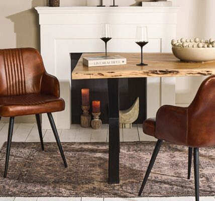 A contemporary dining room featuring a rustic wooden table with live edges, flanked by two brown leather chairs with black metal legs. The table is set against a muted beige wall with a white fireplace mantel. On the table, there's a decorative bowl filled with spheres, a book titled 'Live Beautiful', and two black-stemmed wine glasses. The scene is completed by a textured dark rug underneath and a large potted plant to the left, adding a touch of greenery to the warm-toned space.