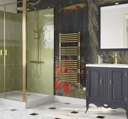 The image features a stylish bathroom with a contemporary design that incorporates brass elements. The focal point is a brushed brass dual fuel towel rail, which stands out against a dark, intricately patterned wallpaper that depicts a lush forest scene with hints of wildlife and florals in deep reds and greens. The shower enclosure showcases a geometric brass frame with clear glass, complementing the towel rail and adding a touch of modern luxury. The vanity unit is a classic dark wood with brass handles, contrasting the white basin above it and the white and black diamond patterned tile floor below. The space is well-lit, and there is a white-framed mirror above the vanity that reflects the natural light coming from the window. The overall aesthetic is one of elegance and warmth, with the brass accents lending a timeless yet trendy feel to the bathroom, exemplifying how brass fixtures can add sophistication to a home.