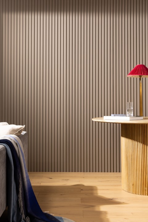 A modern room with beige slatted walls, a sofa with a blue throw, wood flooring, a round table, and a red-shaded lamp casting warm light.