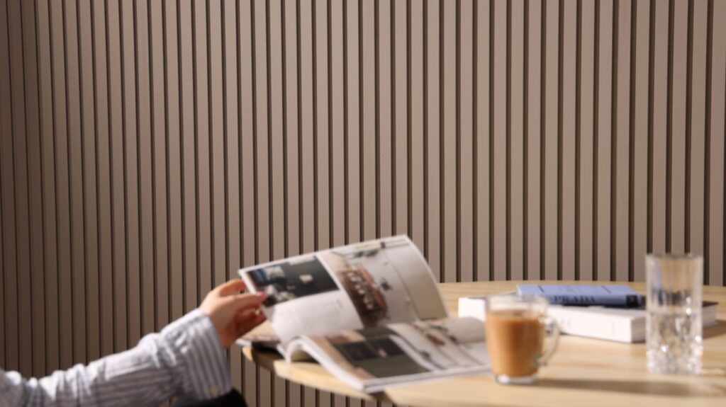 A person reads a magazine at a table. It is an indoor space with a contemporary design, characterized by a slatted wall panelling in the background which adds a textured look to the room. On the table, there's a cup of what might be coffee or tea, a glass of water, and a closed book or journal with a pen on top,