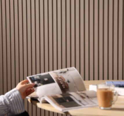 A person reads a magazine at a table. It is an indoor space with a contemporary design, characterized by a slatted wall panelling in the background which adds a textured look to the room. On the table, there's a cup of what might be coffee or tea, a glass of water, and a closed book or journal with a pen on top,