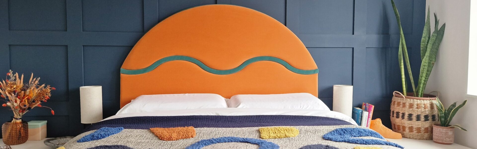 The image shows a bedroom with a contemporary design. The focal point is a large, striking headboard upholstered in a bold orange fabric with a curvilinear teal detail, suggesting a modern and playful style. The headboard sits against a dark blue paneled wall, adding a sense of depth and texture to the room. The bed is covered with a textured blanket featuring abstract shapes in various colors, such as blue, yellow, and orange, which complements the headboard. On either side of the bed are simple, modern white bedside tables, each with a beige lamp, providing a neutral balance to the vibrant colors. There are also decorative elements like a dried flower arrangement in a textured vase, books, and a wicker basket containing green potted plants, enhancing the room's cozy and lived-in feel. The overall composition suggests a well-curated space that blends bold colors and geometric shapes with organic textures.