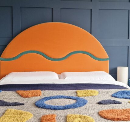 The image shows a bedroom with a contemporary design. The focal point is a large, striking headboard upholstered in a bold orange fabric with a curvilinear teal detail, suggesting a modern and playful style. The headboard sits against a dark blue paneled wall, adding a sense of depth and texture to the room. The bed is covered with a textured blanket featuring abstract shapes in various colors, such as blue, yellow, and orange, which complements the headboard. On either side of the bed are simple, modern white bedside tables, each with a beige lamp, providing a neutral balance to the vibrant colors. There are also decorative elements like a dried flower arrangement in a textured vase, books, and a wicker basket containing green potted plants, enhancing the room's cozy and lived-in feel. The overall composition suggests a well-curated space that blends bold colors and geometric shapes with organic textures.