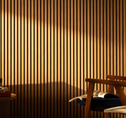 The image shows a living room with a light colored wooden slat wall on the far back wall. There is a gray couch along the left wall, with a light colored wooden end table next to it. A woven basket sits on the floor next to the end table. On the right side of the image is a light colored wooden armchair with a brown leather cushion. A small round coffee table sits in the center of the room, in front of the couch. A black lamp with a white shade sits on the coffee table.