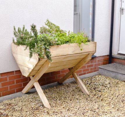 A wooden planter filled with a variety of colourful flowers and plants sits on a gravel surface next to a red brick wall.
