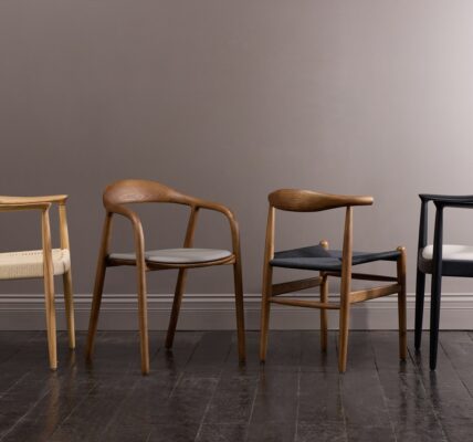 Four wooden chairs with brown upholstery in a row on a light wooden floor. The chairs are against a plain light wall.