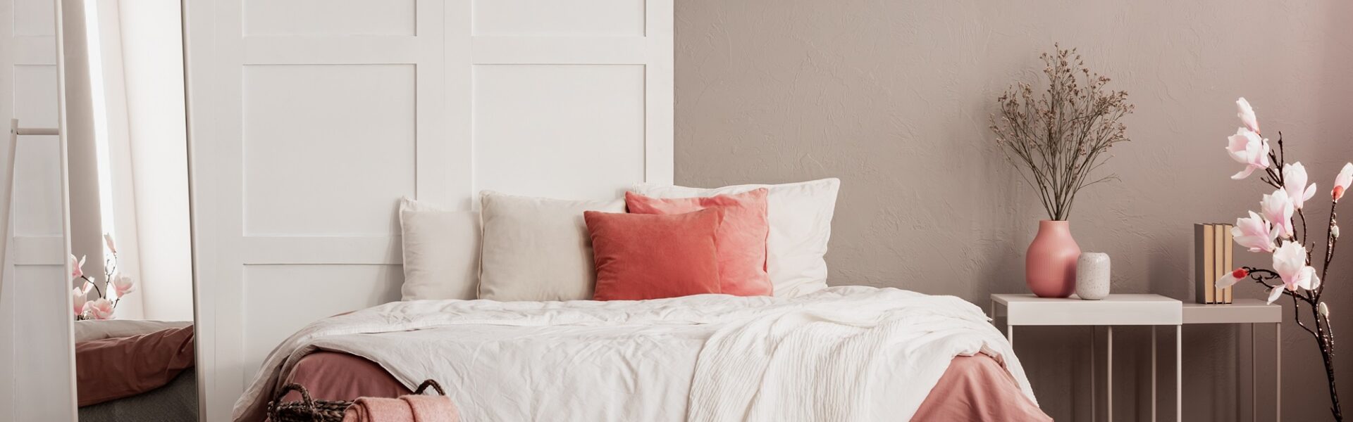 A cosy bedroom with a wooden bed frame, a nightstand with a lamp on top, and a large rectangular mirror hanging on the wall above the nightstand. The bedding is white and the walls are cream coloured.