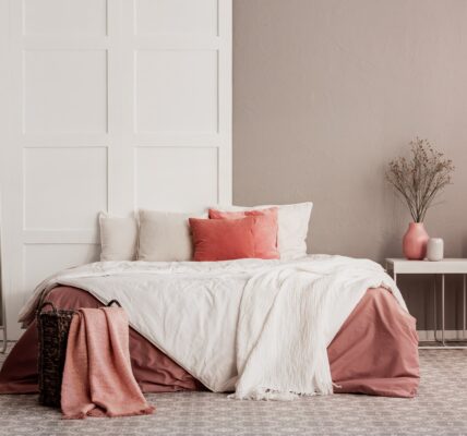 A cosy bedroom with a wooden bed frame, a nightstand with a lamp on top, and a large rectangular mirror hanging on the wall above the nightstand. The bedding is white and the walls are cream coloured.