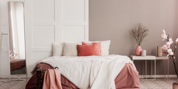 A cosy bedroom with a wooden bed frame, a nightstand with a lamp on top, and a large rectangular mirror hanging on the wall above the nightstand. The bedding is white and the walls are cream coloured.