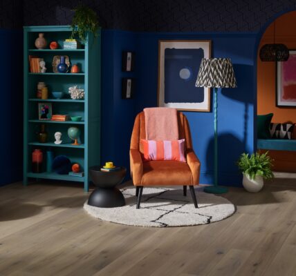 A cosy living room with a wooden armchair upholstered in light fabric sitting on a colourful rug. A wooden bookshelf filled with books is next to the armchair. A table lamp with a white shade sits on a small table beside the armchair.