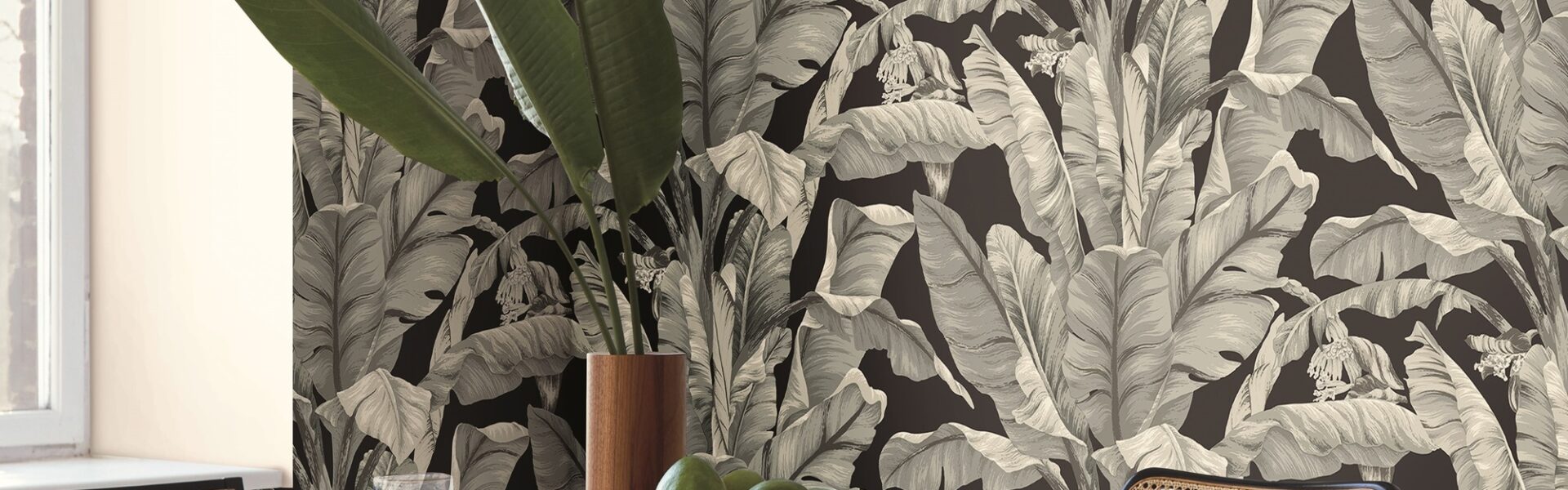 A round wooden dining table with four chairs around it. In the centre of the table is a white ceramic vase filled with bananas. The table sits on a light-coloured rug in a dining room with dark green patterned wallpaper.