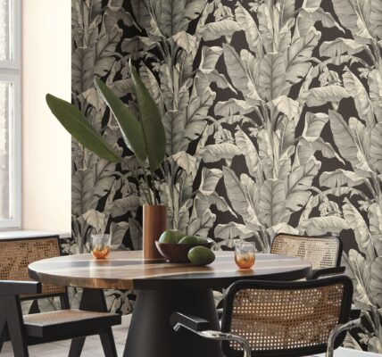 A round wooden dining table with four chairs around it. In the centre of the table is a white ceramic vase filled with bananas. The table sits on a light-coloured rug in a dining room with dark green patterned wallpaper.