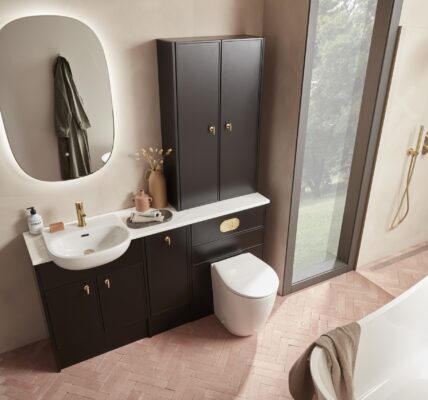 A bright and clean bathroom with a sink, toilet, and mirror. The bathroom has white walls and a tiled floor. There is a window on the left wall. In the foreground, there is a small plant on the sink counter.