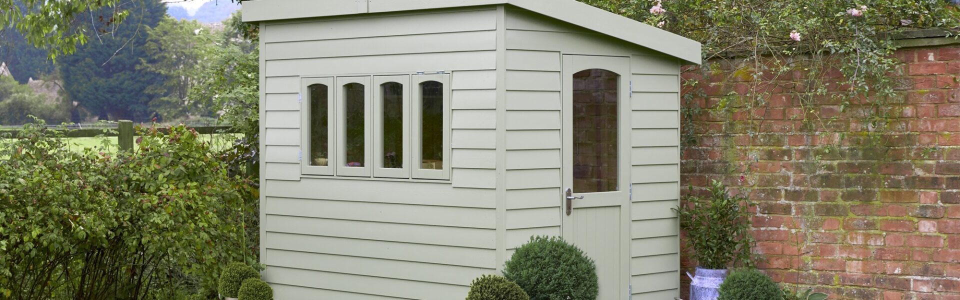 A wooden shed in a garden with a red brick wall behind it. The shed has a pent roof and a door. There are climbing plants on the left-hand side of the wall.