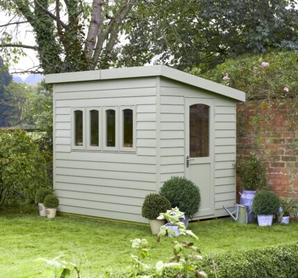 A wooden shed in a garden with a red brick wall behind it. The shed has a pent roof and a door. There are climbing plants on the left-hand side of the wall.