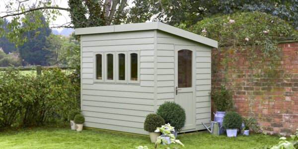 A wooden shed in a garden with a red brick wall behind it. The shed has a pent roof and a door. There are climbing plants on the left-hand side of the wall.
