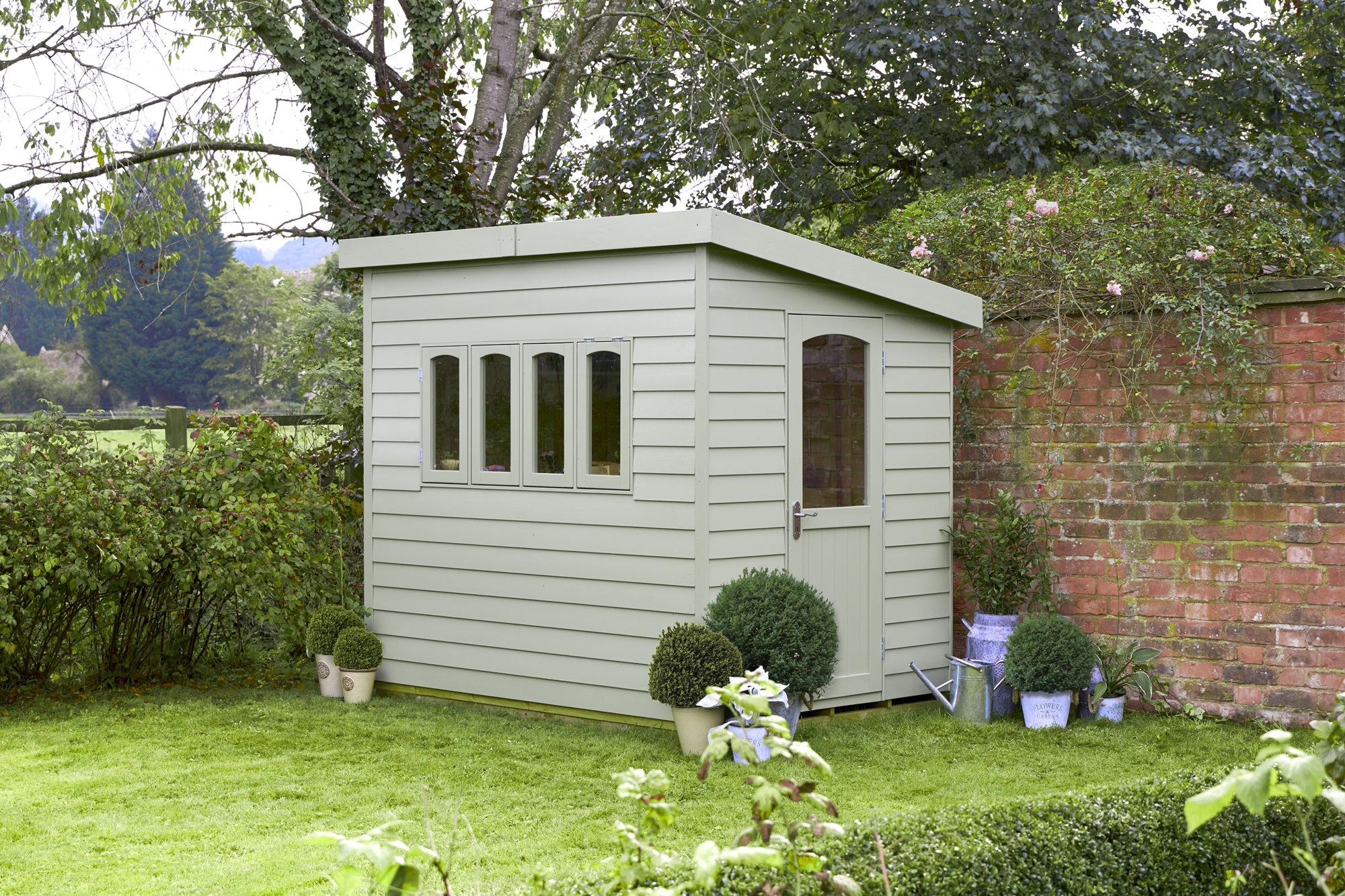 A wooden shed in a garden with a red brick wall behind it. The shed has a pent roof and a door. There are climbing plants on the left-hand side of the wall.