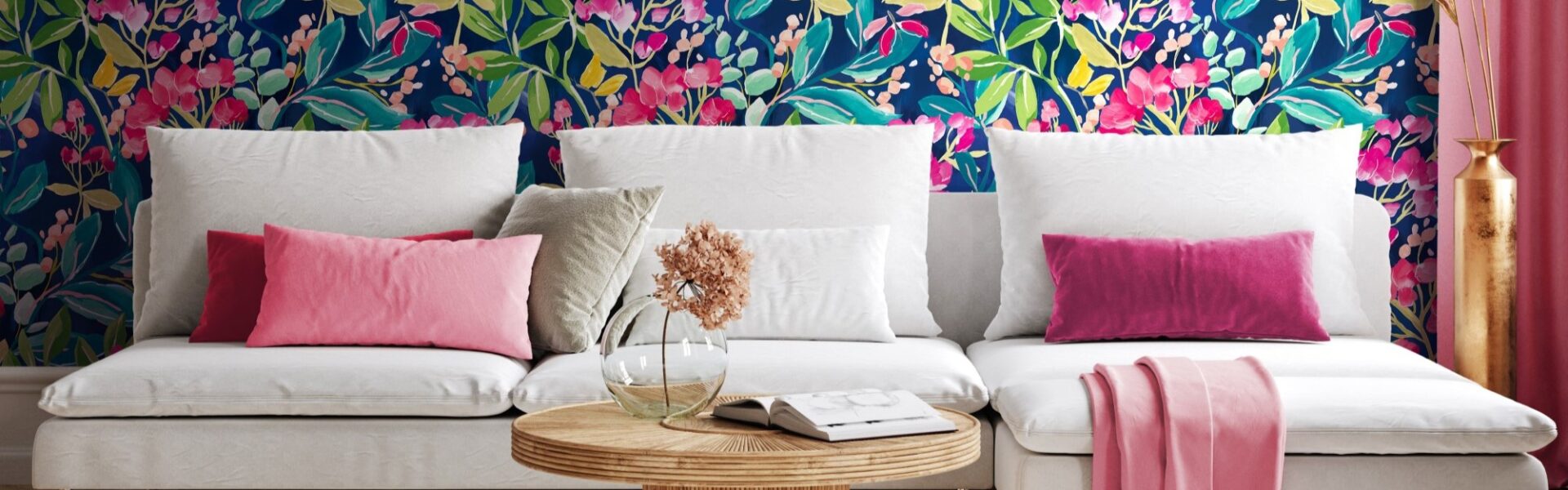 Living room with colourful floral wallpaper on one wall and a pink couch with a round coffee table in front of it. There is a white lamp on the coffee table and a green plant behind the couch.