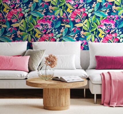 Living room with colourful floral wallpaper on one wall and a pink couch with a round coffee table in front of it. There is a white lamp on the coffee table and a green plant behind the couch.
