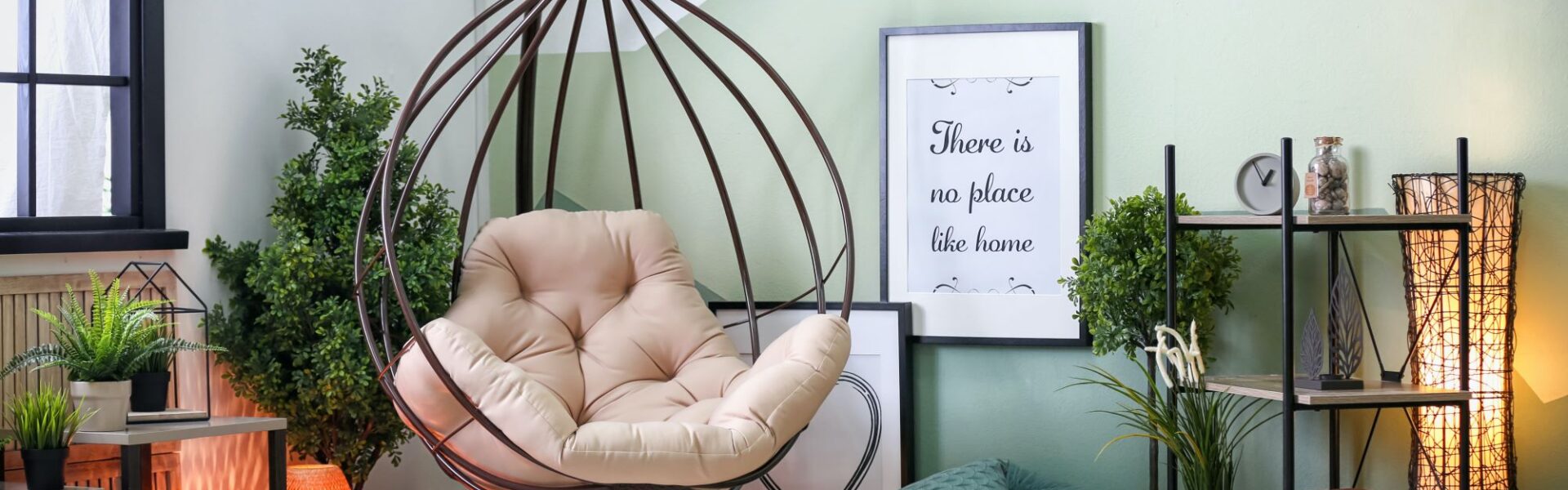 A cosy living room in sage green with a hanging chair in the corner. The chair is made of woven rope and has a cream-coloured cushion. There is a small side table next to the chair with a cup of coffee and a book on it.
