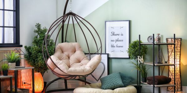 A cosy living room in sage green with a hanging chair in the corner. The chair is made of woven rope and has a cream-coloured cushion. There is a small side table next to the chair with a cup of coffee and a book on it.