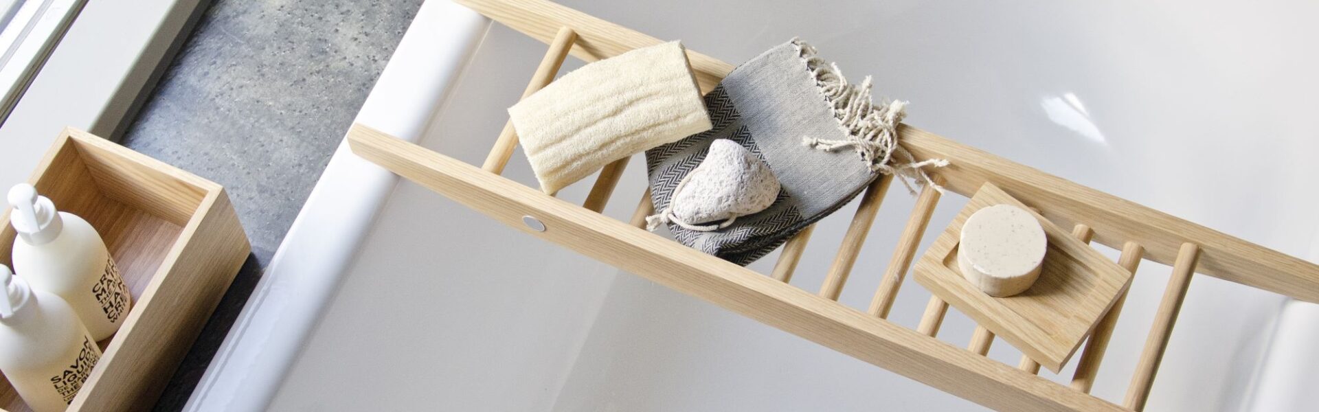 The image shows a Wireworks Bath Bridge in a bathtub. The bath bridge is made of natural oak and has a ladder-like design. It is placed across the bathtub and holds a variety of bath accessories, including a towel, soap, and a loofah. The bathtub is white and has a modern design.