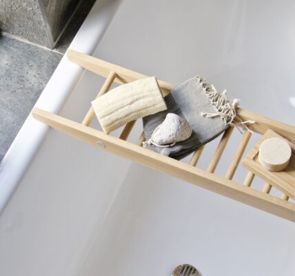 The image shows a Wireworks Bath Bridge in a bathtub. The bath bridge is made of natural oak and has a ladder-like design. It is placed across the bathtub and holds a variety of bath accessories, including a towel, soap, and a loofah. The bathtub is white and has a modern design.