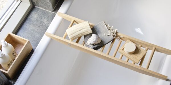 The image shows a Wireworks Bath Bridge in a bathtub. The bath bridge is made of natural oak and has a ladder-like design. It is placed across the bathtub and holds a variety of bath accessories, including a towel, soap, and a loofah. The bathtub is white and has a modern design.