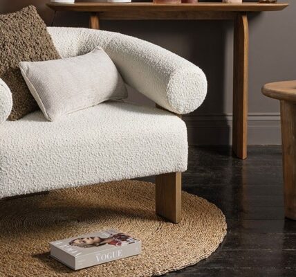 A cosy and stylish living space with a prominent white armchair taking centre stage. The chair has a unique curved design with rolled armrests and is upholstered in a soft, textured fabric. It sits on a round woven rug and is complemented by a wooden side table, a console table, and a collection of decorative items, including pillows, a glass, and books.