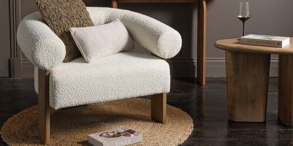A cosy and stylish living space with a prominent white armchair taking centre stage. The chair has a unique curved design with rolled armrests and is upholstered in a soft, textured fabric. It sits on a round woven rug and is complemented by a wooden side table, a console table, and a collection of decorative items, including pillows, a glass, and books.