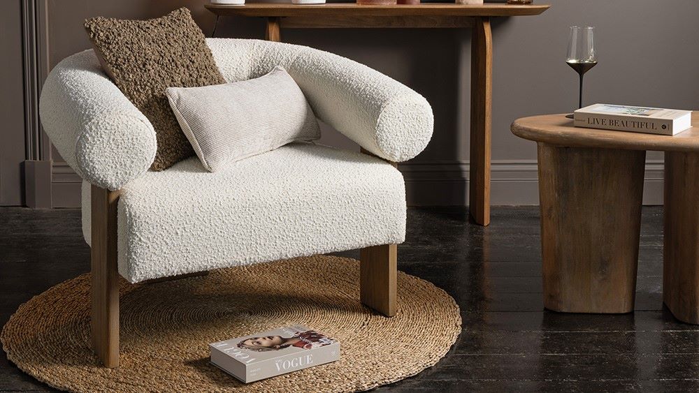 A cosy and stylish living space with a prominent white armchair taking centre stage. The chair has a unique curved design with rolled armrests and is upholstered in a soft, textured fabric. It sits on a round woven rug and is complemented by a wooden side table, a console table, and a collection of decorative items, including pillows, a glass, and books.