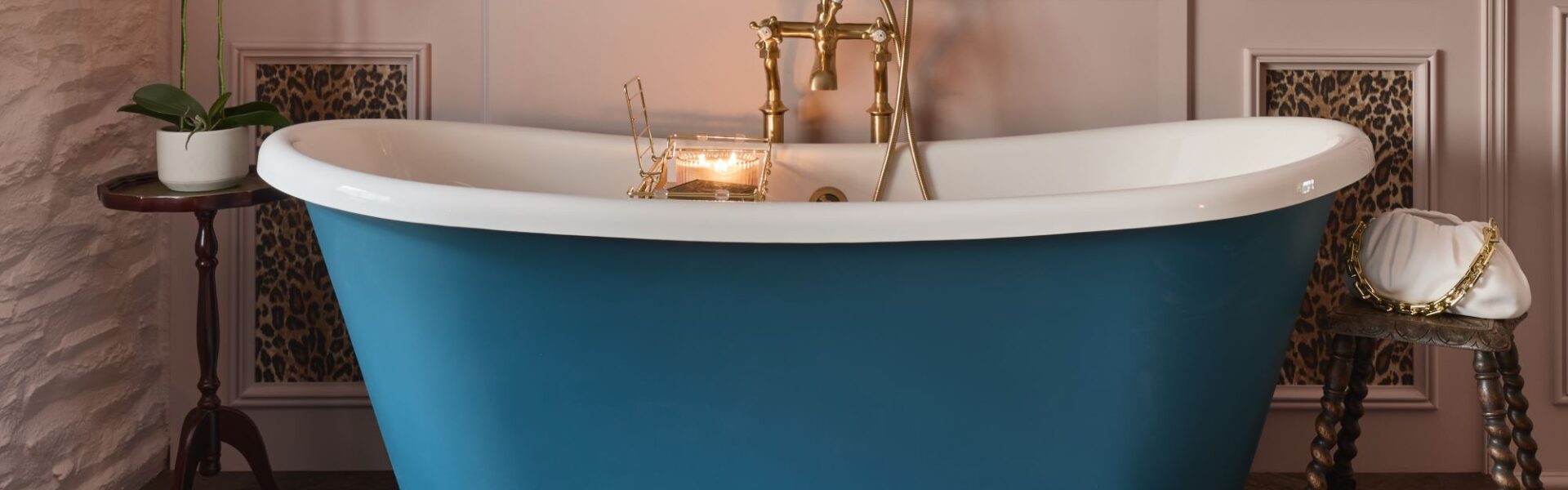 A freestanding bath with a roll top design. It has a white interior and a blue exterior. The bath is sitting on a carpeted floor in front of a wall with leopard print wallpaper. There is a gold faucet on the side of the bath.