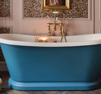 A freestanding bath with a roll top design. It has a white interior and a blue exterior. The bath is sitting on a carpeted floor in front of a wall with leopard print wallpaper. There is a gold faucet on the side of the bath.