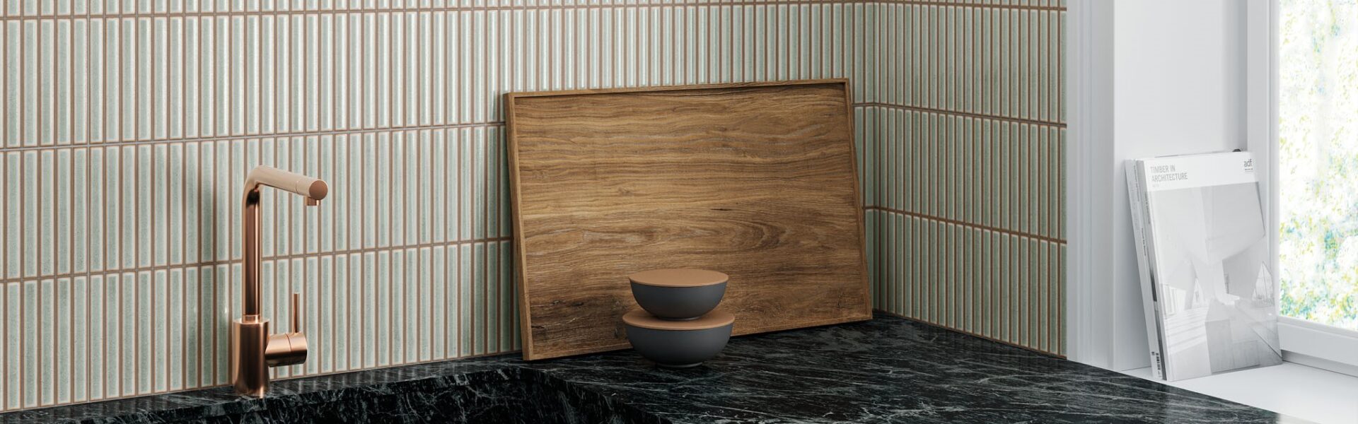 A kitchen countertop with a black marble worktop and a dark green cupboard below. A copper-coloured faucet is mounted on the countertop. There is a wooden cutting board and two bowls on the countertop. The wall behind the countertop is tiled with white tiles with vertical grooves.