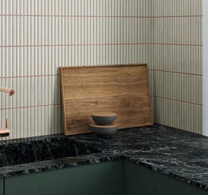 A kitchen countertop with a black marble worktop and a dark green cupboard below. A copper-coloured faucet is mounted on the countertop. There is a wooden cutting board and two bowls on the countertop. The wall behind the countertop is tiled with white tiles with vertical grooves.