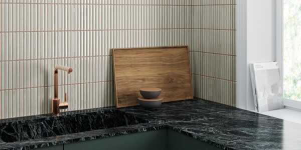 A kitchen countertop with a black marble worktop and a dark green cupboard below. A copper-coloured faucet is mounted on the countertop. There is a wooden cutting board and two bowls on the countertop. The wall behind the countertop is tiled with white tiles with vertical grooves.