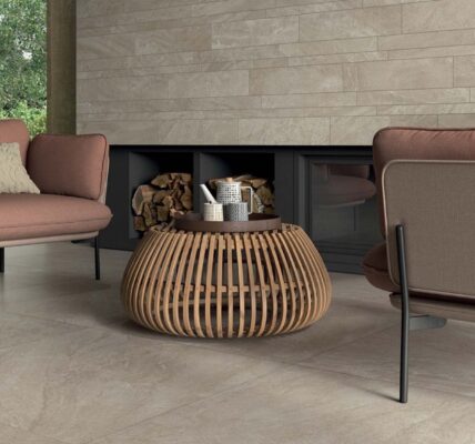 The image shows a spacious and modern living room with two comfortable armchairs facing each other. The chairs have a reddish-brown fabric covering and a black metal frame. A round coffee table with a woven design sits between them. The floor is covered with large, light-coloured tiles, and the walls are adorned with light grey tiles arranged in a brick pattern.