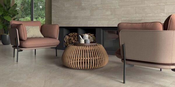 The image shows a spacious and modern living room with two comfortable armchairs facing each other. The chairs have a reddish-brown fabric covering and a black metal frame. A round coffee table with a woven design sits between them. The floor is covered with large, light-coloured tiles, and the walls are adorned with light grey tiles arranged in a brick pattern.