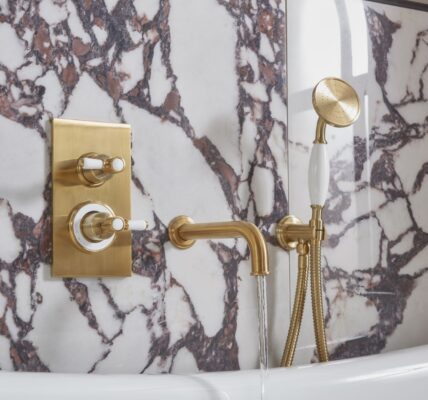 The image shows a luxurious bathroom with a Keswick Dual Function Concealed Shower System. The shower system has a brushed brass finish and features a thermostatic mixer valve with two ceramic grip levers for controlling temperature and flow. The shower head is a large, round overhead shower, and there is also a handheld shower with a flexible hose. The bathroom is also decorated with beautiful marble tiles and a freestanding bathtub.