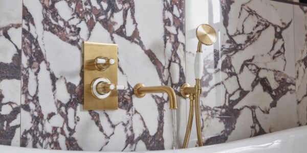 The image shows a luxurious bathroom with a Keswick Dual Function Concealed Shower System. The shower system has a brushed brass finish and features a thermostatic mixer valve with two ceramic grip levers for controlling temperature and flow. The shower head is a large, round overhead shower, and there is also a handheld shower with a flexible hose. The bathroom is also decorated with beautiful marble tiles and a freestanding bathtub.