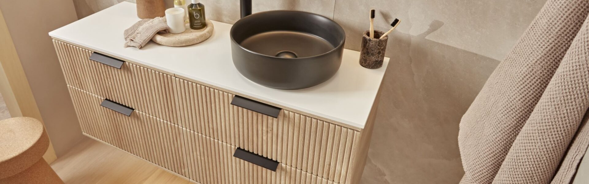 A modern bathroom vanity with a floating wooden cabinet and a black ceramic countertop. A round black sink sits on the countertop, with a black tap and soap dispenser next to it. A fluffy white towel hangs on the wall behind the vanity.
