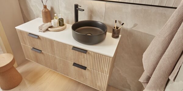 A modern bathroom vanity with a floating wooden cabinet and a black ceramic countertop. A round black sink sits on the countertop, with a black tap and soap dispenser next to it. A fluffy white towel hangs on the wall behind the vanity.