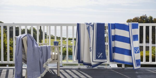 Blue and white striped blankets and throws draped over a white wooden balcony railing, with a blue blanket on a chair overlooking a coastal view.