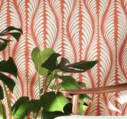 A cosy armchair sits in front of a wall covered in orange and white patterned wallpaper. A large houseplant with lush green leaves stands beside the chair.