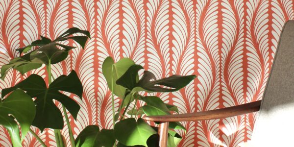 A cosy armchair sits in front of a wall covered in orange and white patterned wallpaper. A large houseplant with lush green leaves stands beside the chair.